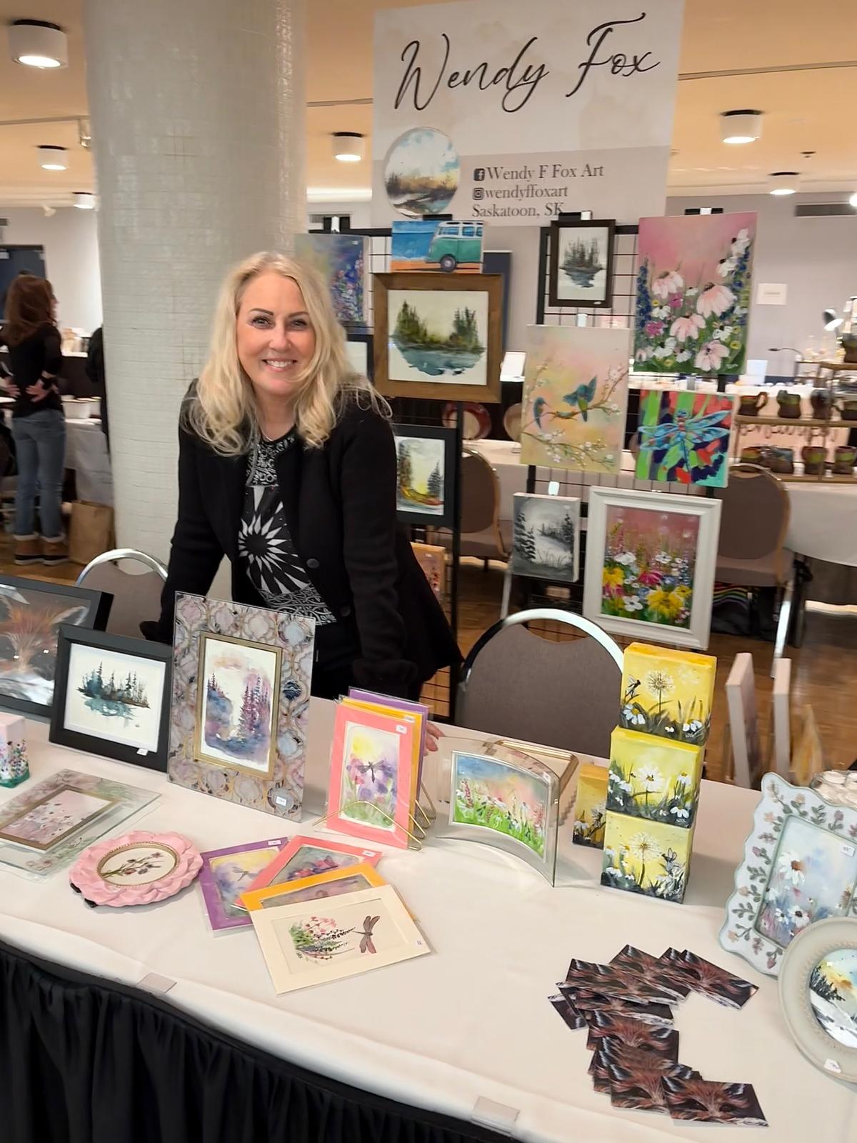 A woman stands behind a craft fair booth displaying handmade art prints, framed cards, and decorative pieces. A sign on the wall behind her reads Wendy Fox Art & Design.